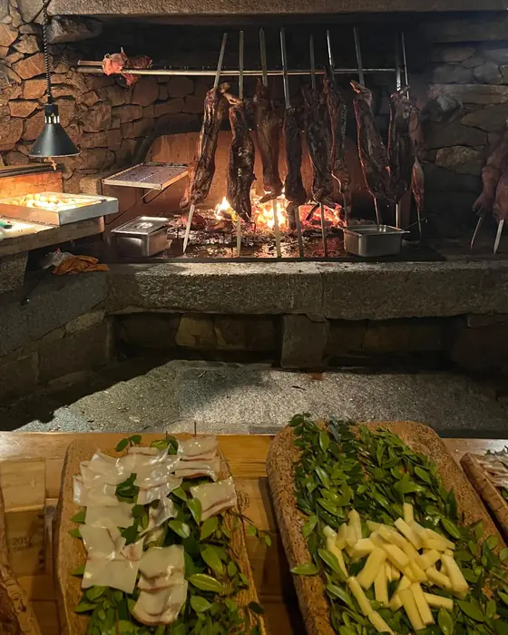 Suckling pig roasting on spits over an open fire in a Sardinian stone hearth, with cured meats and cheese on cork boards